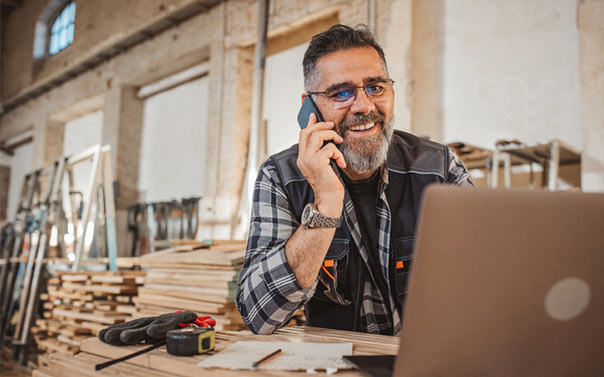Carpenter in wood workshop on phone and laptop